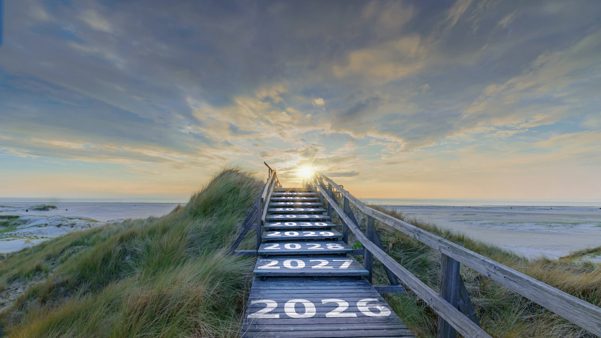 The image shows a wooden staircase leading through dunes to the beach, running towards a sunrise or sunset. The years from 2026 to 2035 are painted large and white on the individual steps, as if symbolising the path to the future. At the end of the staircase, where the light is strongest, the journey seems to lead into the coming years, while the wide sky and calm sea in the background convey a peaceful, expansive mood.