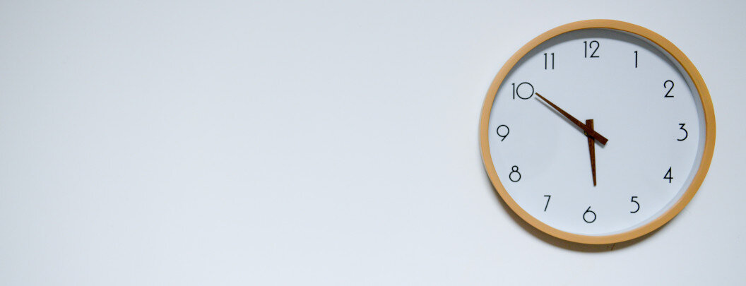 The image shows an analogue wall clock with a yellow frame hanging on a white wall. The time is approximately 6:50 p.m. The clock face has a simple design and the background of the clock and the wall are white, giving the image a very minimalist look. There is a lot of empty space on the left, while the clock is located on the right side of the image.